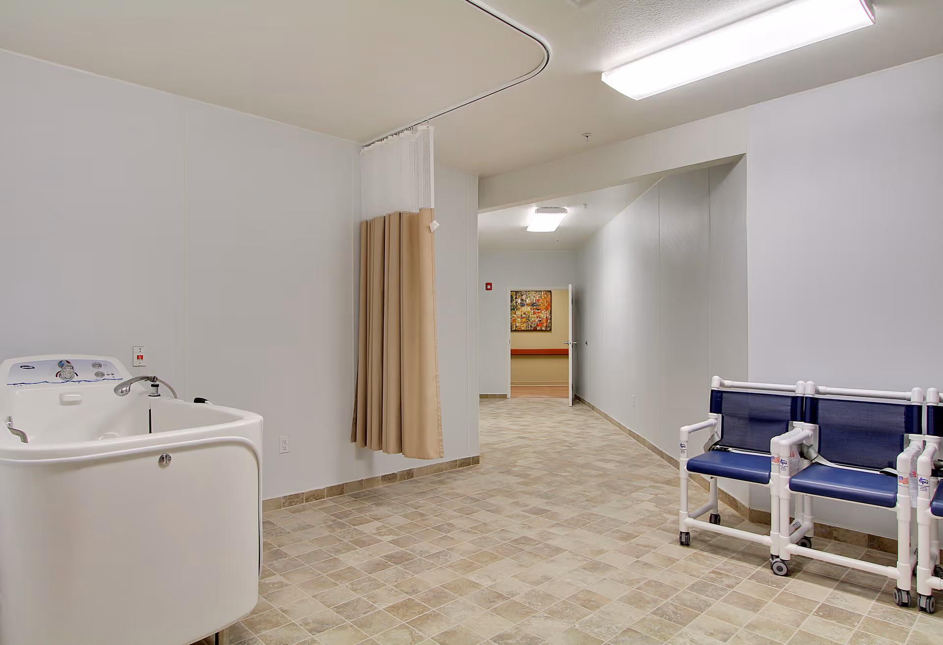 A clean, tiled room with a white bathing tub on the left side and a beige privacy curtain hanging from a ceiling track. On the right side, there are three blue and white chairs with wheels. The room leads to a hallway with a colorful painting on the far wall.