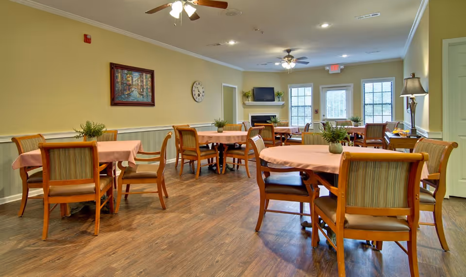 Well-lit communal dining room with round tables covered in pink tablecloths, wooden chairs, ceiling fans, and a TV above a fireplace.