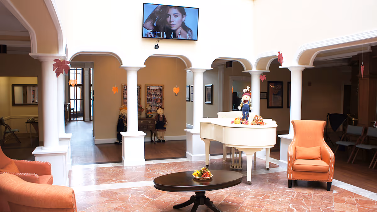 A bright and spacious common area with white columns and arches, featuring a white grand piano decorated with autumn-themed items. Two orange upholstered armchairs flank a dark oval coffee table with a small centerpiece. A flat-screen TV is mounted high on the wall, and autumn leaf decorations hang from the ceiling. The floor is a mix of marble and wood, and the space has a warm, inviting atmosphere.