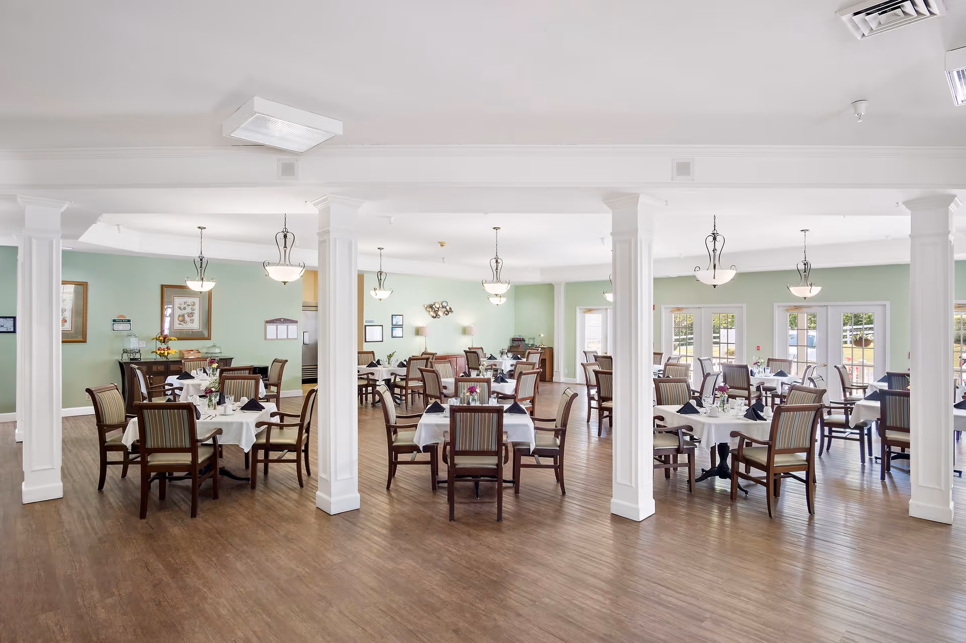 A spacious dining room with multiple tables covered in white tablecloths, each set with plates, glasses, and napkins. The room has wooden floors, light green walls, white columns, and several hanging light fixtures. Large windows and glass doors allow natural light to fill the space.