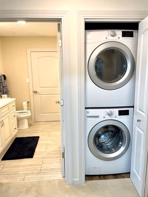 A laundry area with a stacked washer and dryer unit inside a closet with white doors. To the left, there is a bathroom with a toilet, a vanity with a sink, a black rug on the floor, and beige tiled flooring.