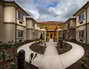 Outdoor courtyard area of a senior living facility with a circular walkway, landscaped plants, benches, and a wooden pergola under a cloudy sky. The building has two stories with beige walls and brown trim.