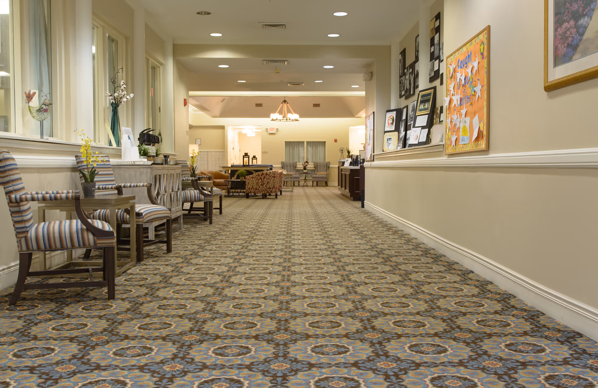 Long carpeted interior hallway with patterned carpeting, chairs and small tables along the left wall and a seating area at the far end.