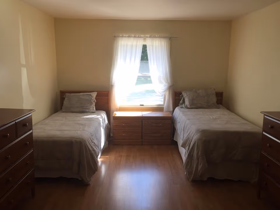 A simple bedroom with two single beds placed on either side of a window with sheer white curtains. Each bed has a pillow and light-colored bedding. Between the beds is a wooden nightstand with drawers. On each side of the room, there is a wooden dresser. The room has light-colored walls and wooden flooring.