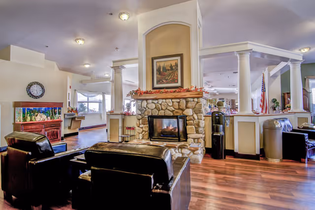 A cozy living room area in a senior living facility featuring a stone fireplace with a painting above it, surrounded by leather armchairs. The room has wooden flooring, white columns, a large fish tank, and an American flag in the background.