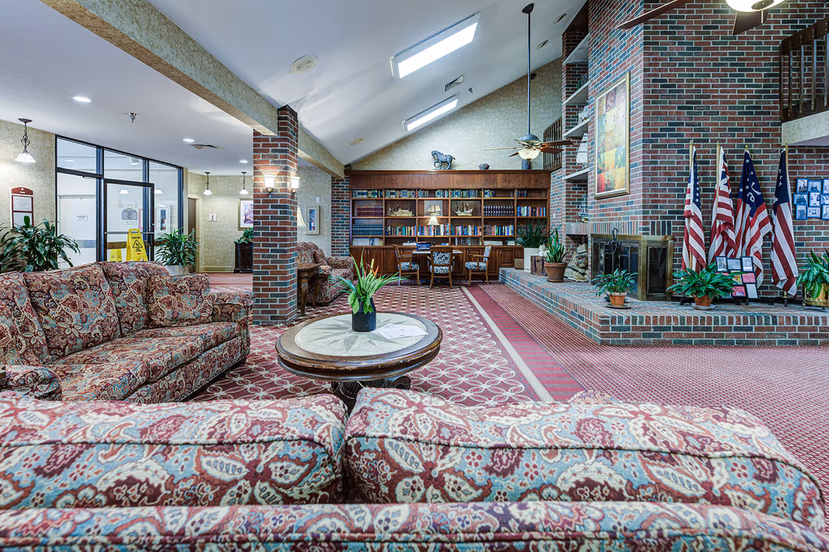 A spacious senior living facility common area with patterned upholstered sofas and a round wooden coffee table with a plant centerpiece. The room features a large brick fireplace adorned with American flags and potted plants. There is a built-in wooden bookshelf filled with books and decorative items, and a table with chairs in front of it. The ceiling is vaulted with skylights and ceiling fans, and the floor is covered with a patterned carpet.