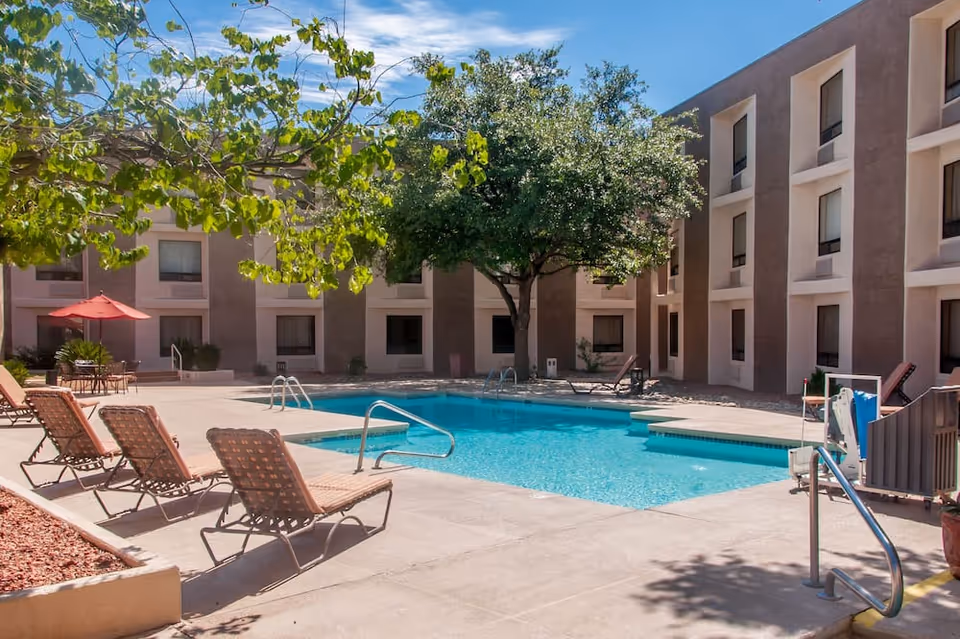 Outdoor swimming pool area at a senior living facility with lounge chairs, a large tree providing shade, and a building with multiple windows surrounding the pool. There is also a red umbrella over a table with chairs in the background.
