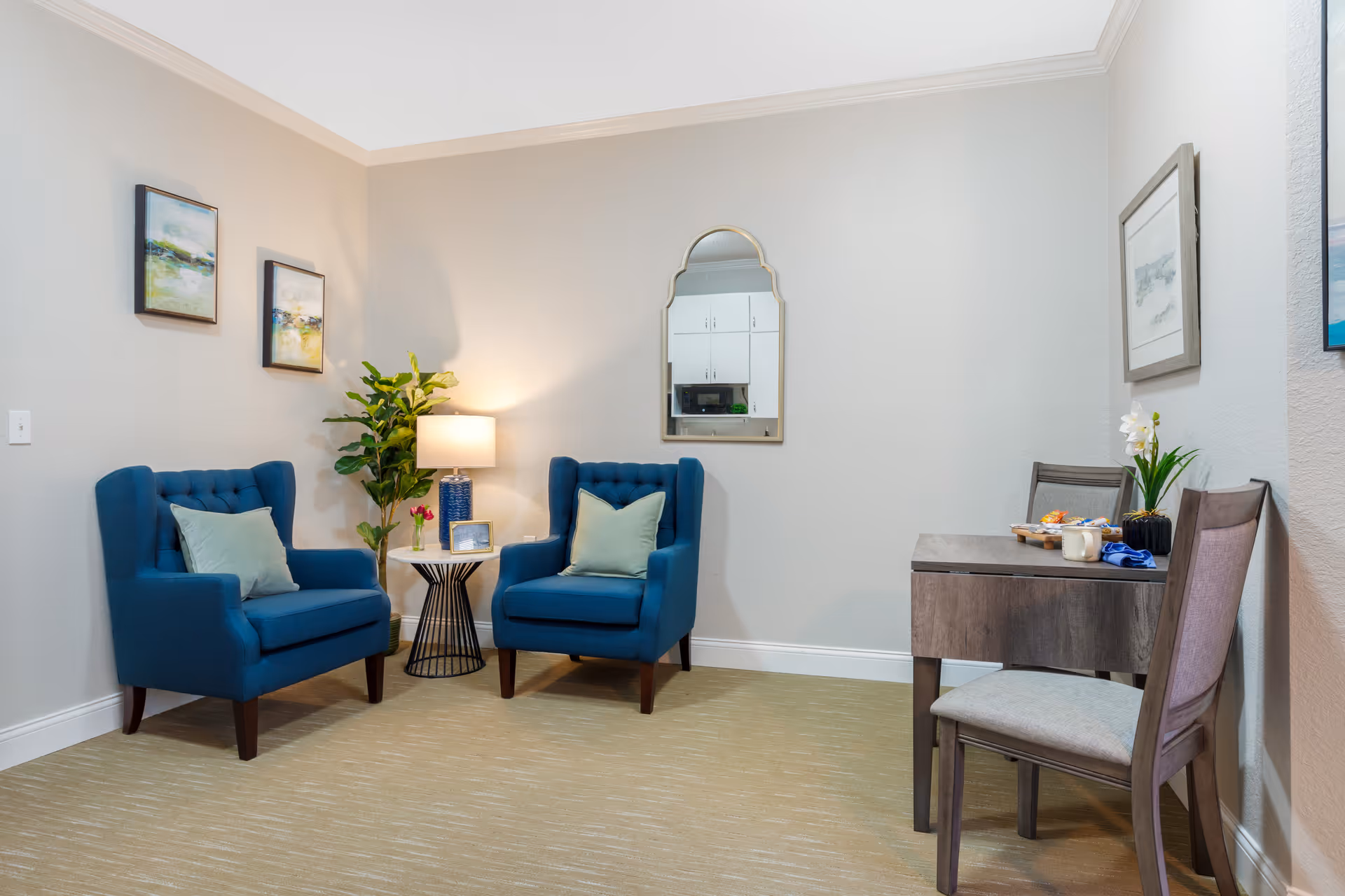 A cozy sitting area with two blue upholstered armchairs with light green pillows, a small round side table with a lamp and a plant, a wall mirror reflecting white cabinets, and a wooden desk with two chairs, a plant, and snacks on top.
