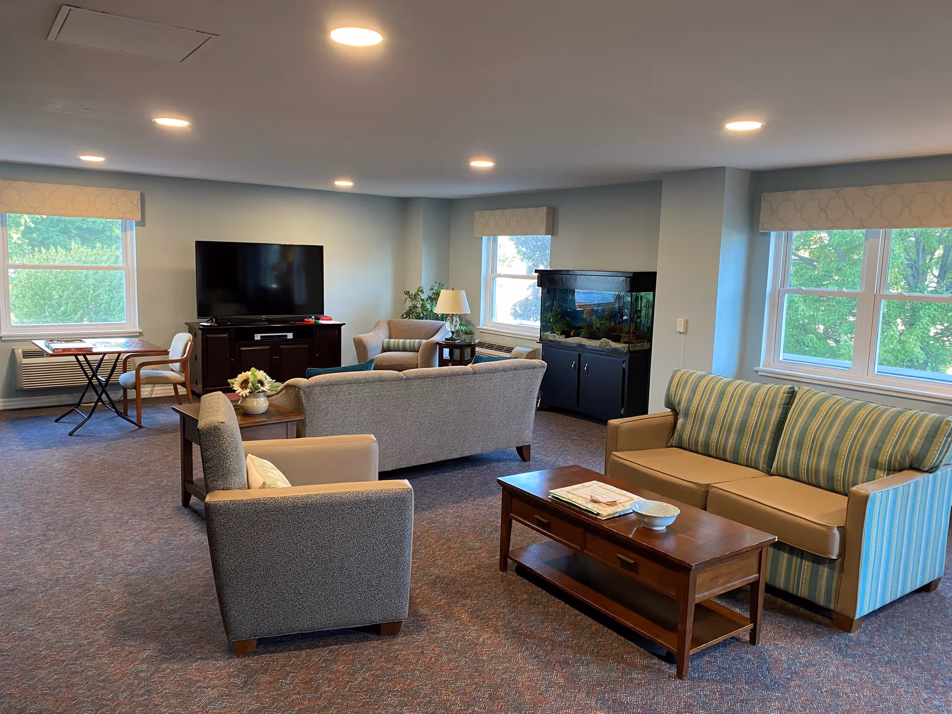 A spacious living room with multiple seating options including a striped sofa, a beige sofa, and two armchairs. There is a wooden coffee table with a bowl and some papers on it. A large flat-screen TV is placed on a dark wooden cabinet against the wall. The room has several windows with light-colored valances, allowing natural light to brighten the space. A large fish tank is positioned near one of the windows. The carpet is patterned in muted colors, and the ceiling has recessed lighting.