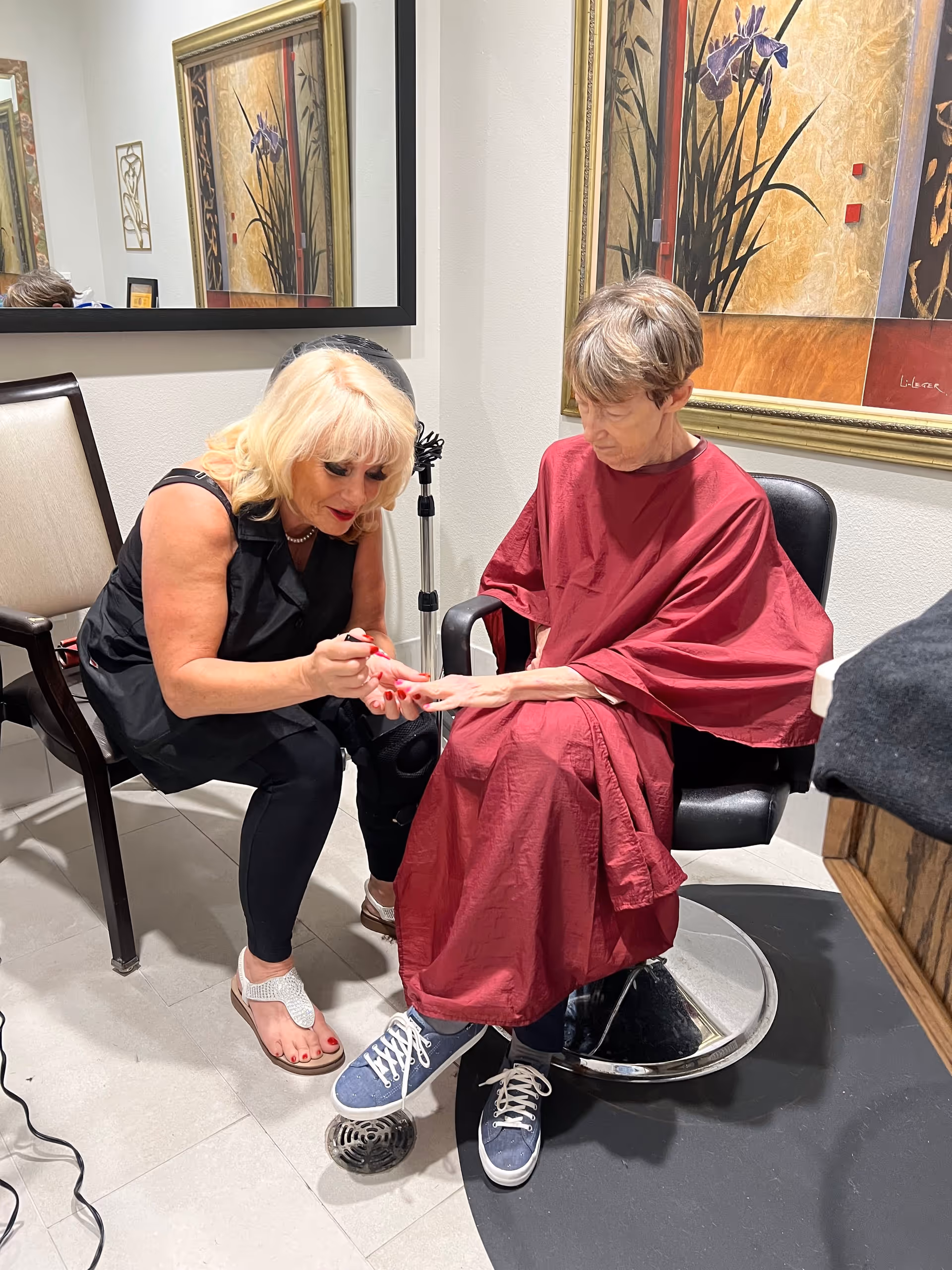 A woman with blonde hair is painting the nails of an elderly woman seated in a salon chair wearing a red cape. The setting appears to be a salon or beauty area with framed artwork on the walls and a large mirror reflecting part of the scene.