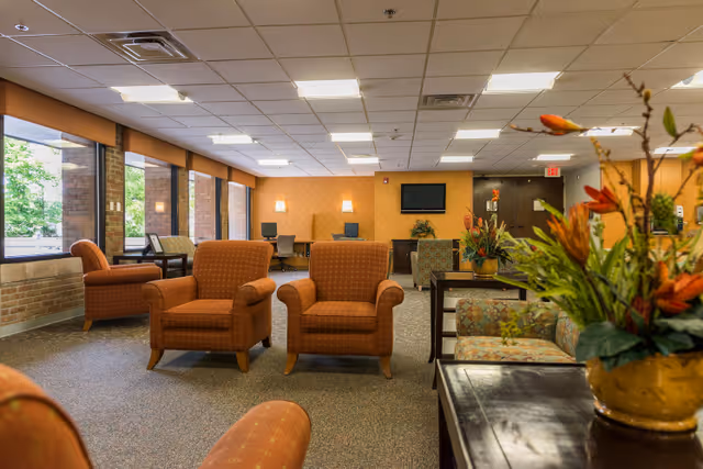 A spacious senior living facility common area with multiple orange upholstered armchairs and patterned sofas arranged around tables. Large windows on the left side let in natural light, and the walls are painted in warm tones. There is a flat-screen TV mounted on the far wall above a cabinet, and decorative plants and flowers add a touch of greenery to the room.