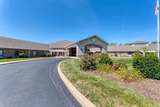 Exterior view of Cedarhurst of Breese facility showing a curved driveway leading to the main entrance with a covered drop-off area. The building is made of brick with a pitched roof, surrounded by well-maintained landscaping including bushes and flowers under a clear blue sky.