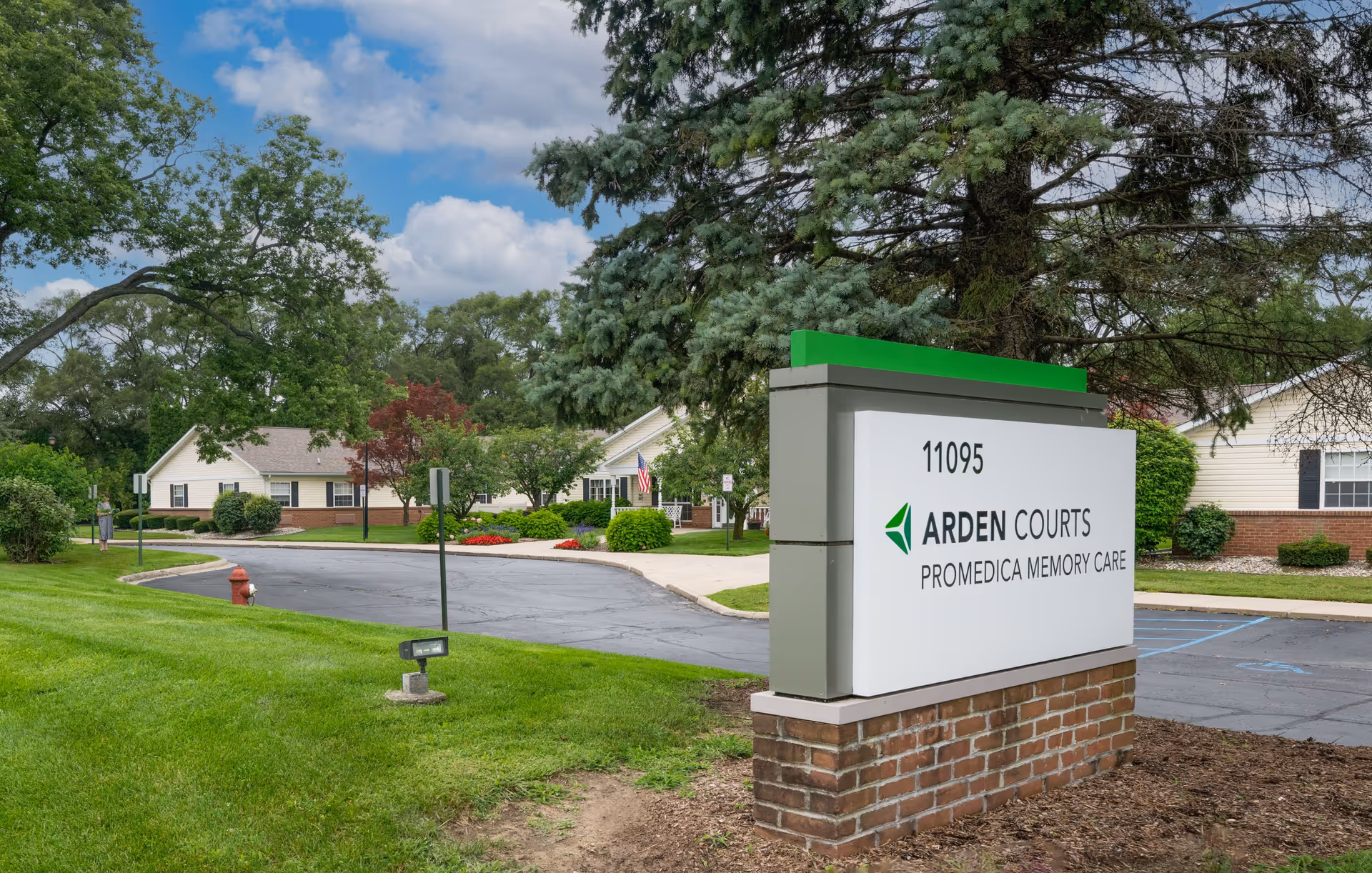 Outdoor view of Arden Courts ProMedica Memory Care Community in Sterling Heights, showing the facility's entrance sign with trees, grass, and buildings in the background under a partly cloudy sky.