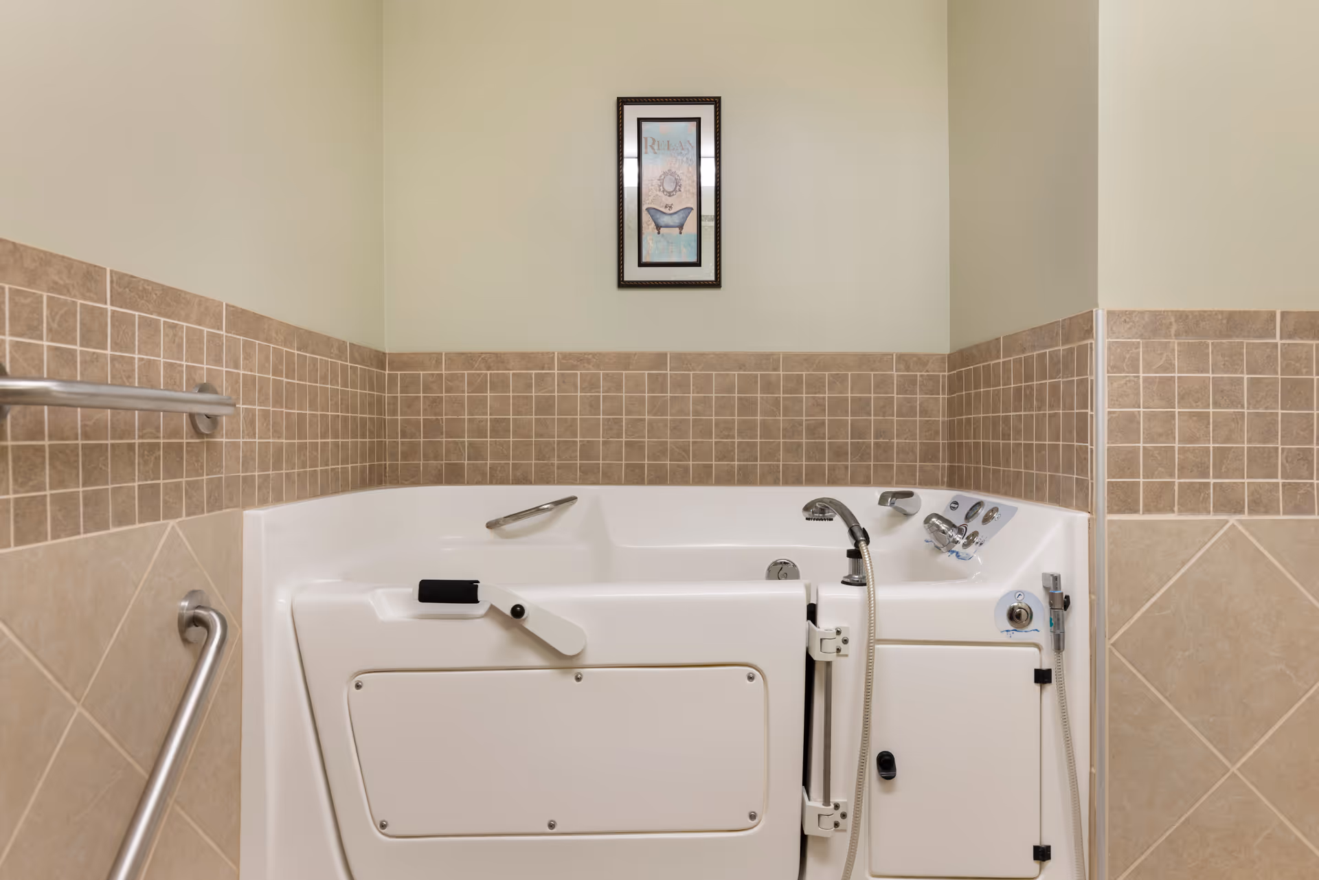 A walk-in bathtub with safety handles and a handheld showerhead, surrounded by beige and brown tiled walls. Above the tub is a framed picture with the word 'RELAX' and an image of a bathtub.