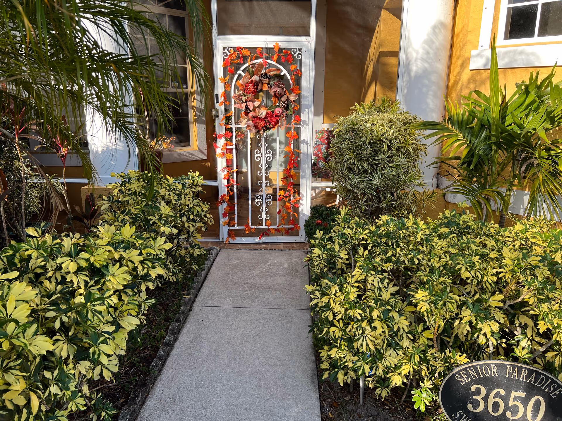 Entrance to a building with a white decorative metal door adorned with a fall-themed wreath and garland. The pathway leading to the door is flanked by lush green and yellow variegated bushes and palm plants. The building exterior is painted yellow with white trim around the windows and door. A black oval sign with gold lettering reads 'Senior Paradise 3650'.
