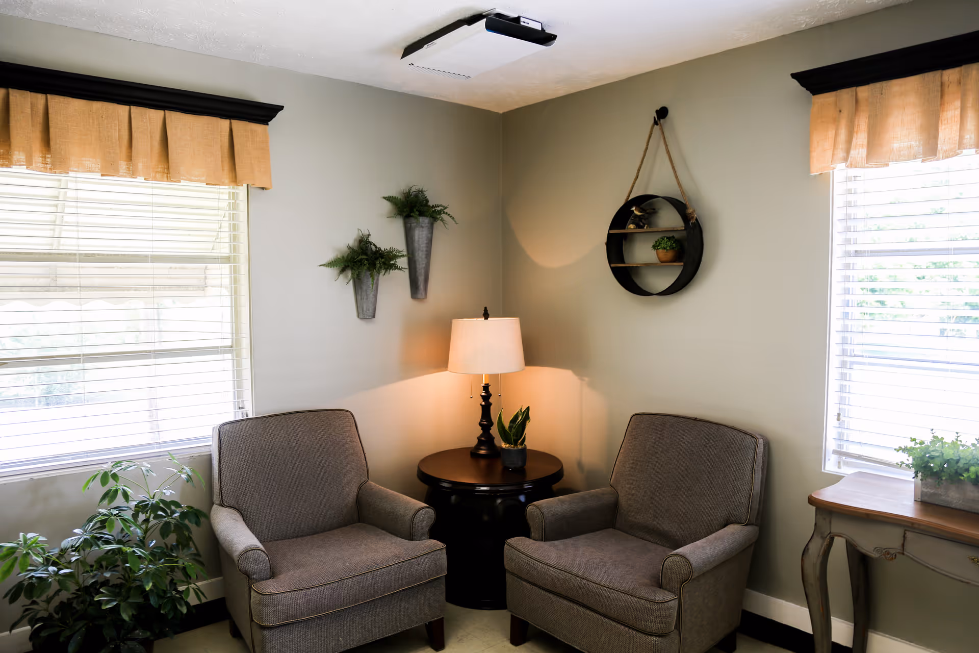 Two upholstered armchairs sit around a round side table with a lamp in a cozy seating nook between two windows and potted plants.