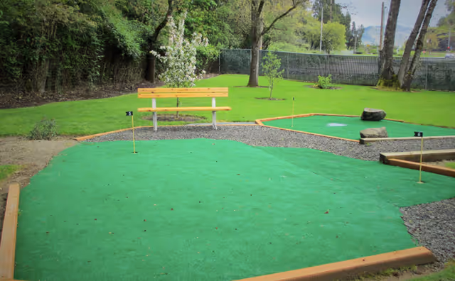 Outdoor artificial putting greens with small flags, a wooden bench, and trees in a grassy yard.