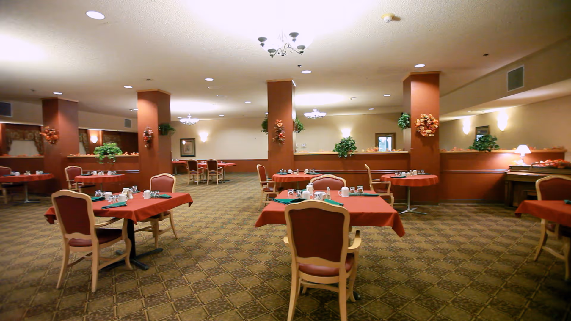 A spacious dining room with multiple tables covered in red tablecloths, each set with cups, napkins, and utensils. The room has patterned carpet flooring, several columns decorated with wreaths, wall sconces, chandeliers, and some green plants placed around. Chairs with red upholstery are arranged around the tables.