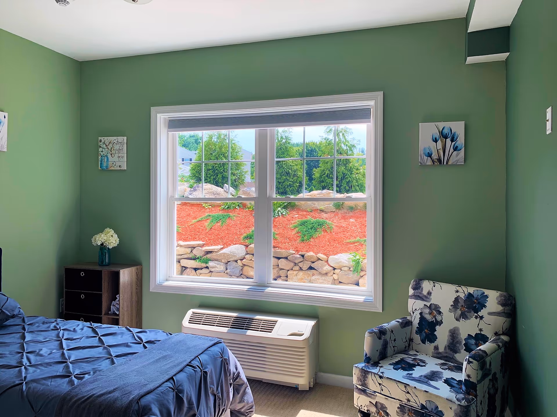 A bedroom with green walls featuring a window with a view of a landscaped garden with rocks and shrubs. The room has a bed with a dark blue quilt, a small wooden nightstand with a vase of white flowers, and a floral-patterned armchair.