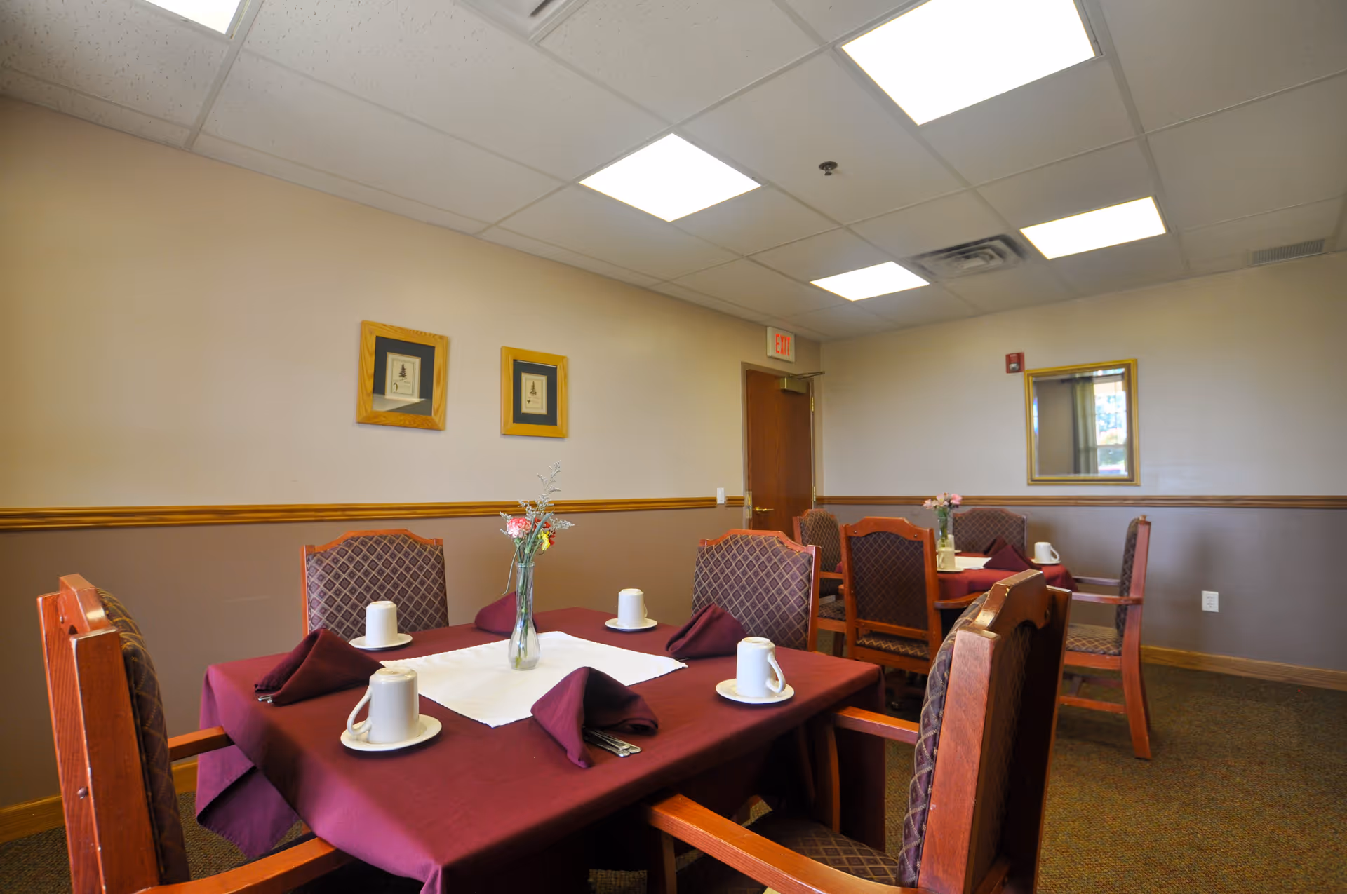 Small dining room with tables set with burgundy tablecloths, chairs, place settings, and ceiling lights.