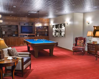 A cozy recreation room with a pool table centered on a red carpet, surrounded by seating, bookshelves, a TV, and wall art.