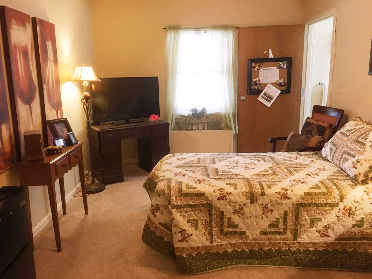 A cozy bedroom with a bed covered in a floral and geometric patterned quilt. There is a wooden desk with a flat-screen TV on it, a floor lamp, and a small table with framed photos and decorative items. A window with sheer curtains is centered on the back wall, and a wooden door with a bulletin board is to the right. A wooden chair with cushions is placed near the door.