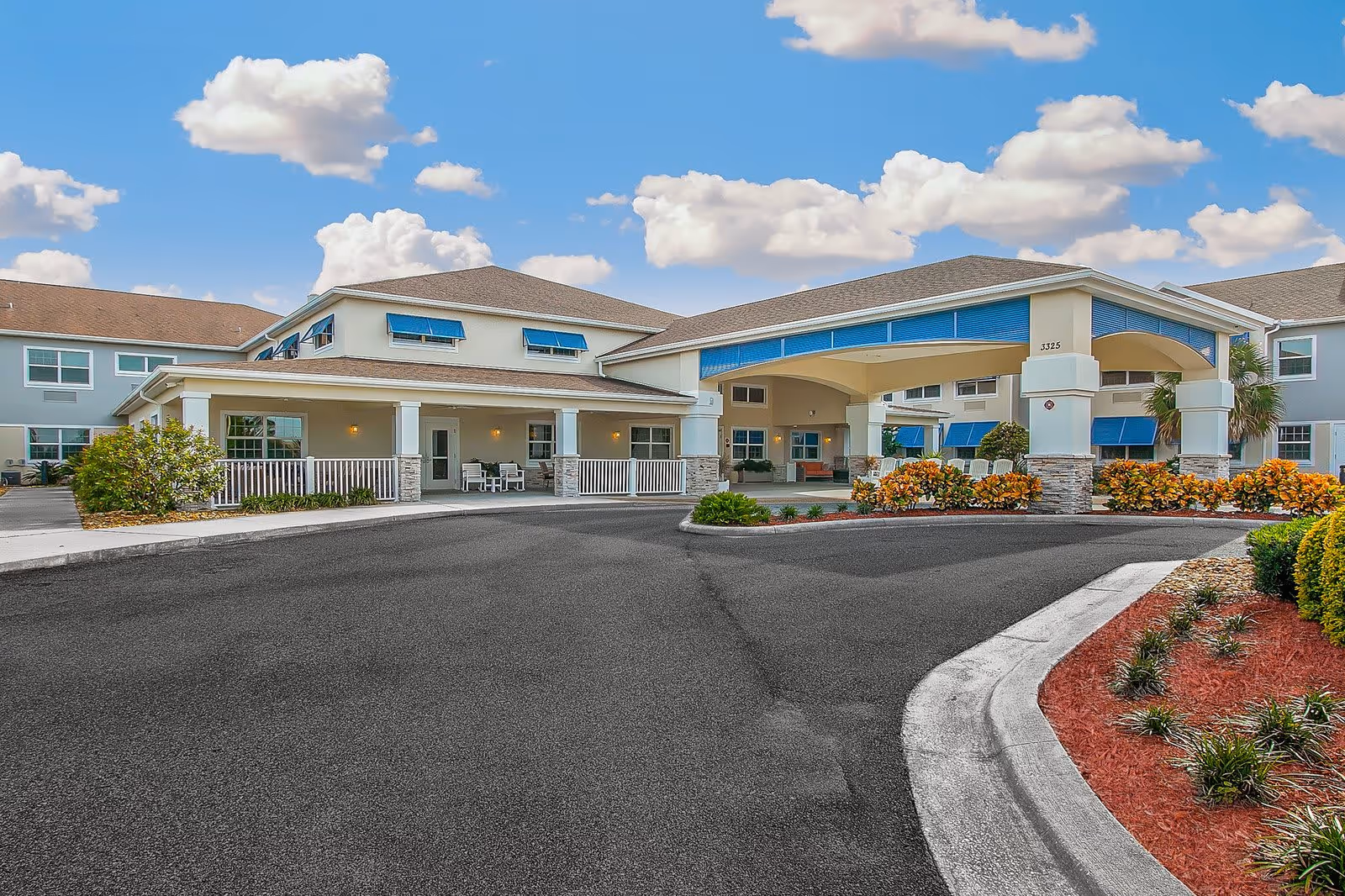 Exterior view of Sonata Viera senior living facility showing the main entrance with a covered drop-off area, surrounded by landscaped bushes and plants under a partly cloudy blue sky.