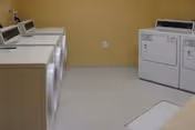 A laundry room with four washing machines on the left side and two dryers on the right side against a beige wall, with a light-colored floor.