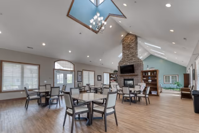 Spacious senior living common area with multiple tables and chairs arranged on a wooden floor. The room features a high vaulted ceiling with skylights and a large stone fireplace with a mounted TV above it. Large windows with blinds allow natural light to fill the space, and there are bookshelves and plants along the walls.