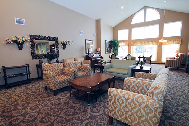 Spacious communal living room with patterned armchairs and sofas arranged around a coffee table beneath large arched windows.
