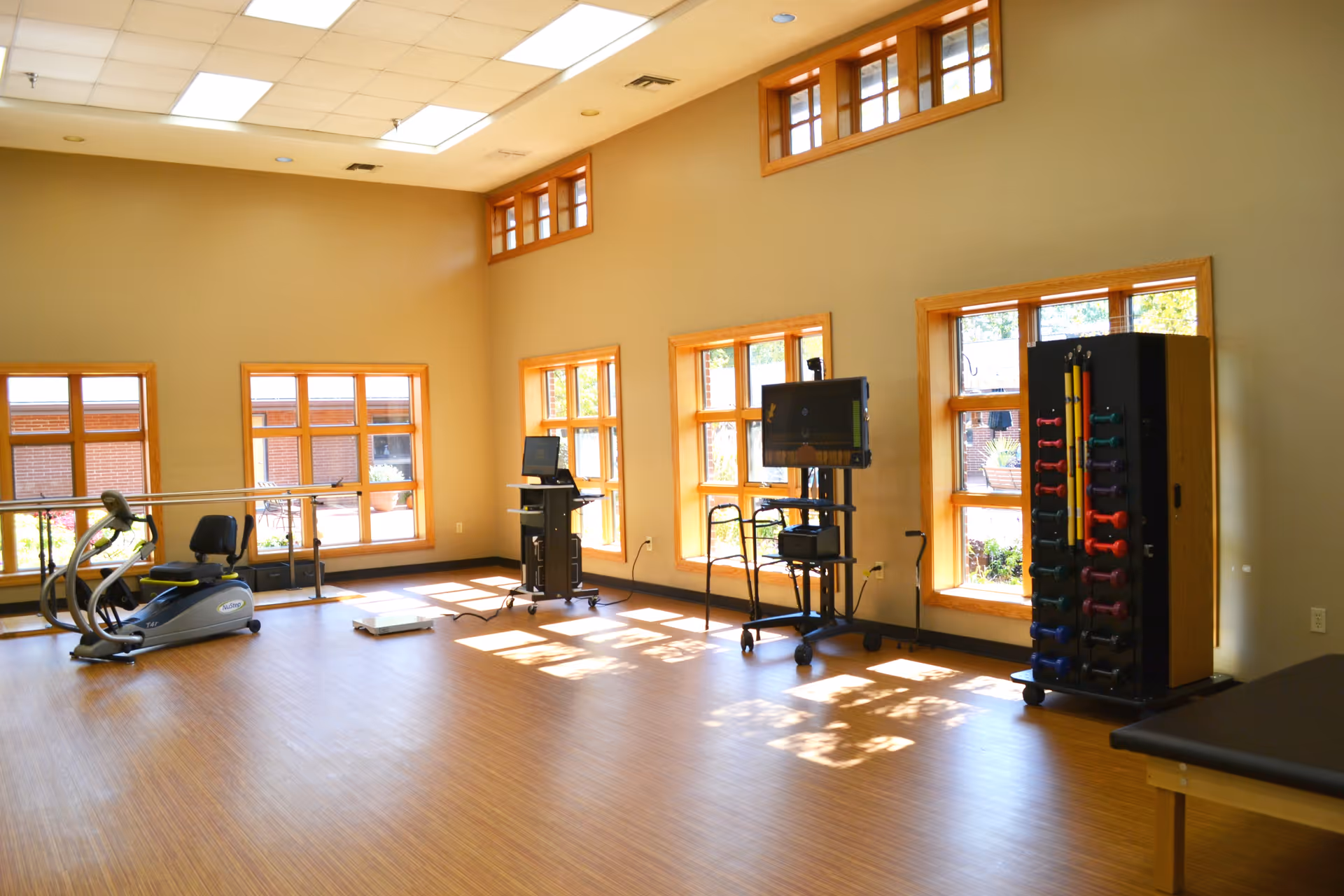 A bright exercise room with wooden floors and multiple large windows letting in natural light. The room contains exercise equipment including a recumbent bike, parallel bars, a scale, a rack of colorful dumbbells, and two computer stations on wheeled carts.