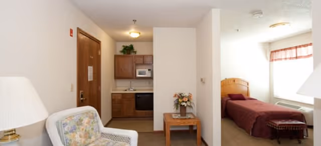 Interior view of a senior living facility room at Brookdale Gallatin showing a small kitchenette with wooden cabinets, a microwave, and a dishwasher. In the foreground, there is a white armchair with floral cushions and a table with a flower arrangement. To the right, a separate sleeping area with a single bed covered in a burgundy blanket is visible next to a window with a valance.