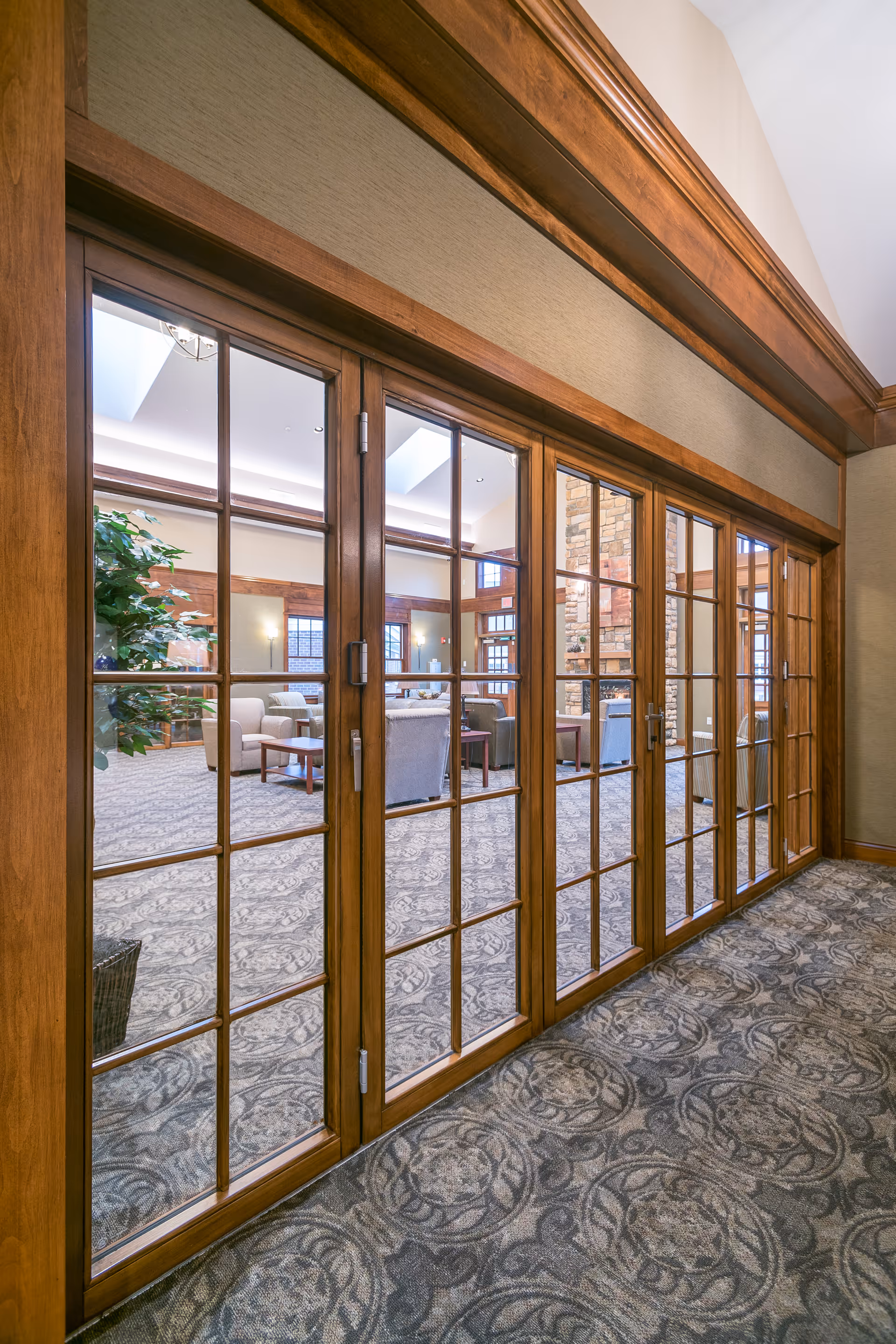 View through large wooden framed glass doors into a spacious living room area with patterned carpet, comfortable armchairs, a coffee table, and a stone fireplace. The room has high ceilings and large windows allowing natural light.
