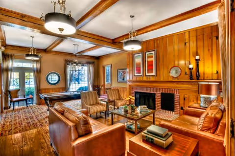 A cozy living room with wooden panel walls and ceiling beams, featuring leather sofas, striped armchairs, a glass coffee table with books and flowers, a brick fireplace, and framed artwork above the mantel. Large windows with curtains let in natural light, and a pool table is visible in the background near a door leading outside.