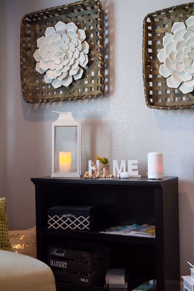 A cozy corner with a black shelf holding decorative items including a white lantern with a candle, wooden letters spelling HOME, and a small plant. Above the shelf are two woven wall hangings with large white flower decorations. A beige cushion with gold text is partially visible on the left.