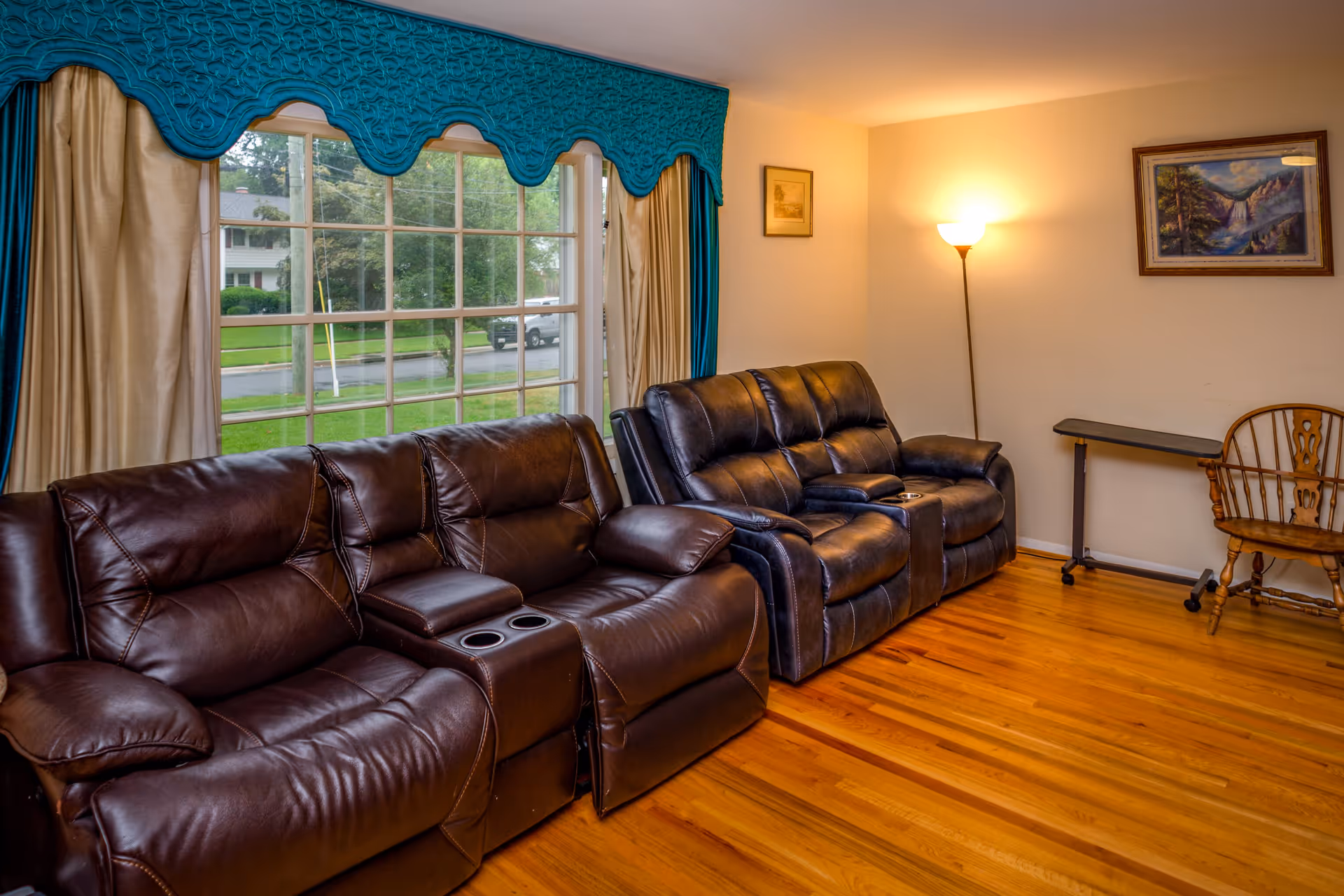 A cozy living room with two dark brown leather reclining sofas placed against a wall with a large window covered by teal and beige curtains. The room has wooden flooring, a floor lamp in the corner, a small table, and a wooden chair. Two framed pictures hang on the walls.