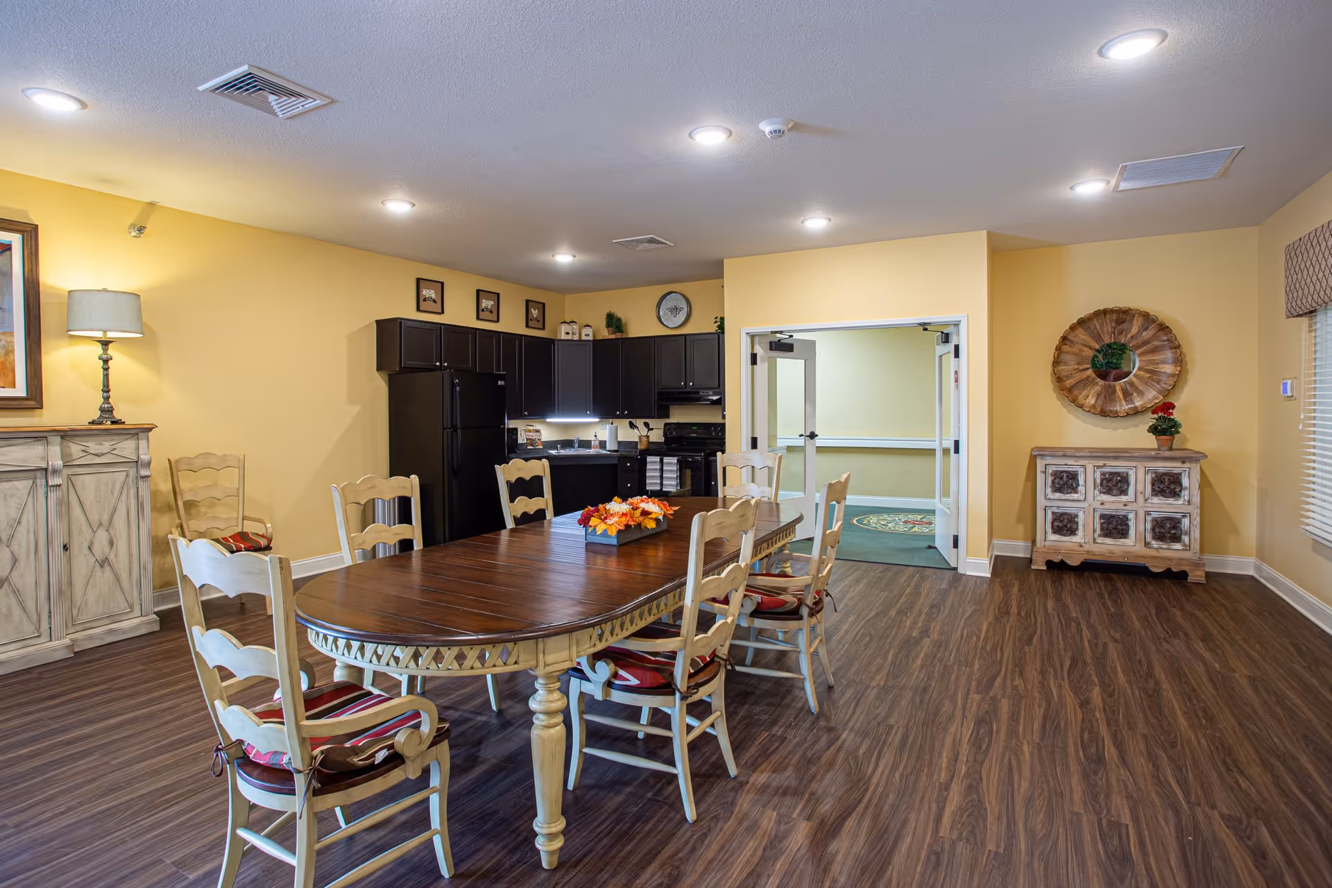 Communal dining room featuring a long wooden table with chairs, a small kitchenette with black cabinets, and warm yellow walls.