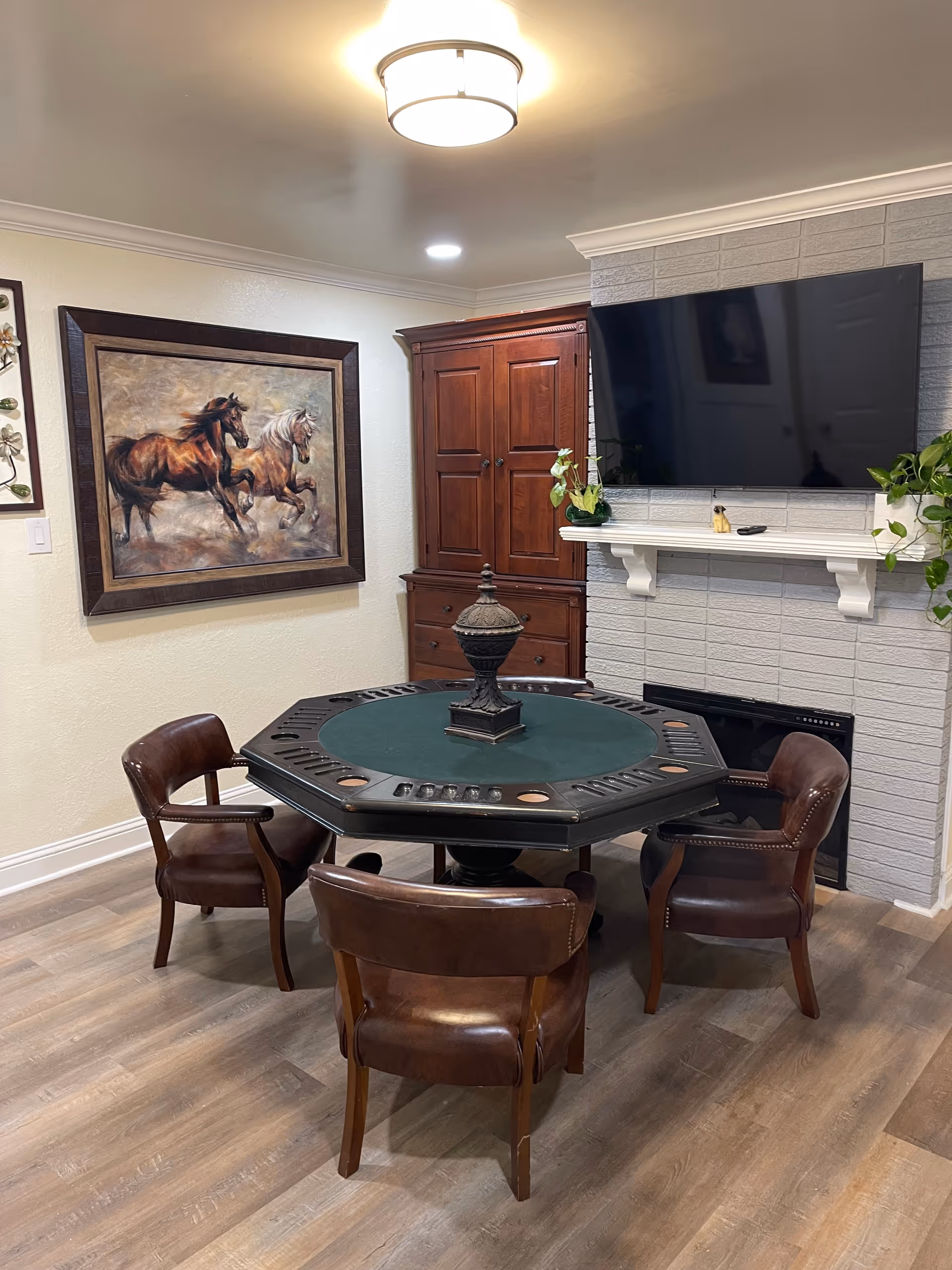 Cozy interior room with a round poker table and four leather chairs, a mounted TV above a fireplace, a wooden cabinet, and a horse painting on the wall.