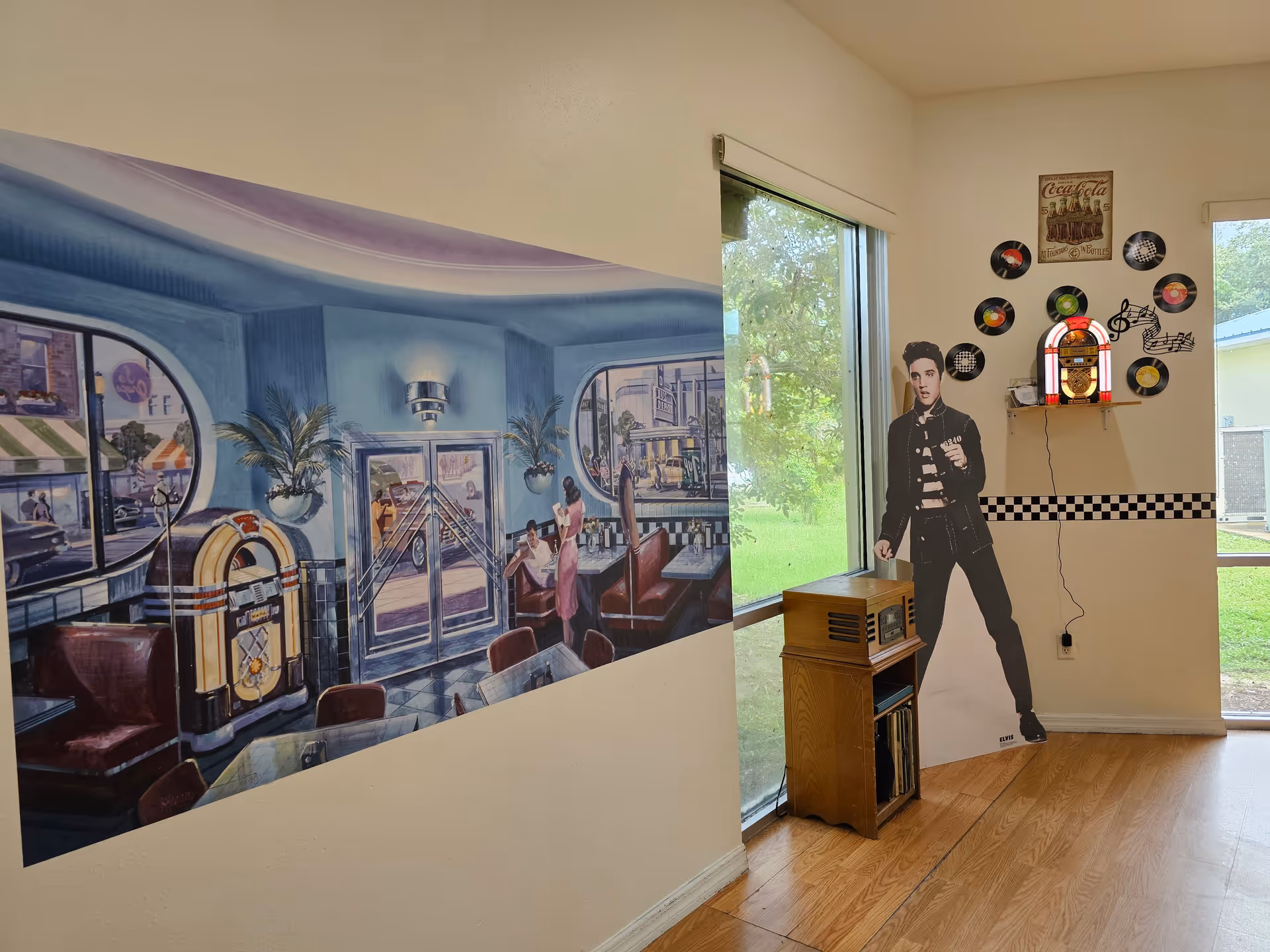 Interior corner of a room with a large mural on one wall depicting a vintage diner scene with a jukebox and people sitting at booths. On the adjacent wall, there is a life-sized cutout of Elvis Presley next to a small wooden cabinet holding a vintage radio and records. Above the cutout, a small shelf holds a retro jukebox and is decorated with vinyl records and a vintage Coca-Cola sign. Large windows show green grass and trees outside.