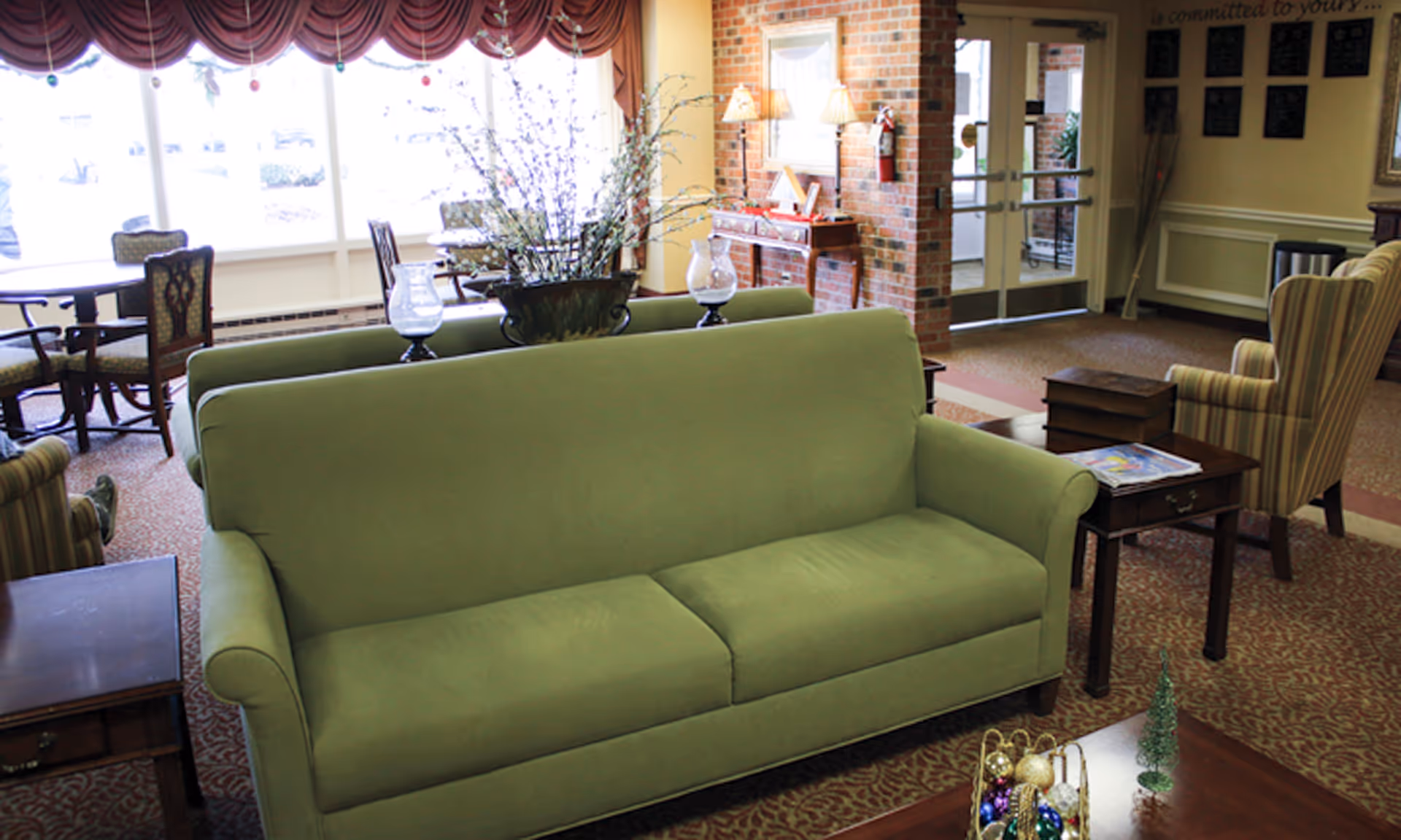 A cozy living room area in a senior living facility featuring a green sofa, striped armchairs, wooden side tables, and a coffee table with holiday decorations. The room has large windows with red valances, a brick wall with a console table and lamps, and a glass door leading outside.