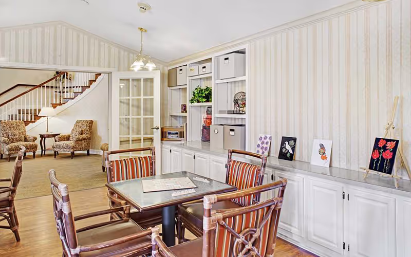 A cozy common area in a senior living facility featuring a square table with four wooden chairs that have striped cushions. The table has a board game set up on it. Behind the table is a built-in white cabinet with shelves holding storage boxes, a plant, and decorative items. On the right side, there are small framed artworks displayed on the countertop. In the background, there is a seating area with two patterned armchairs, a small table with a lamp, and a staircase with a wooden handrail. The walls have light-colored striped wallpaper and the floor is wooden.
