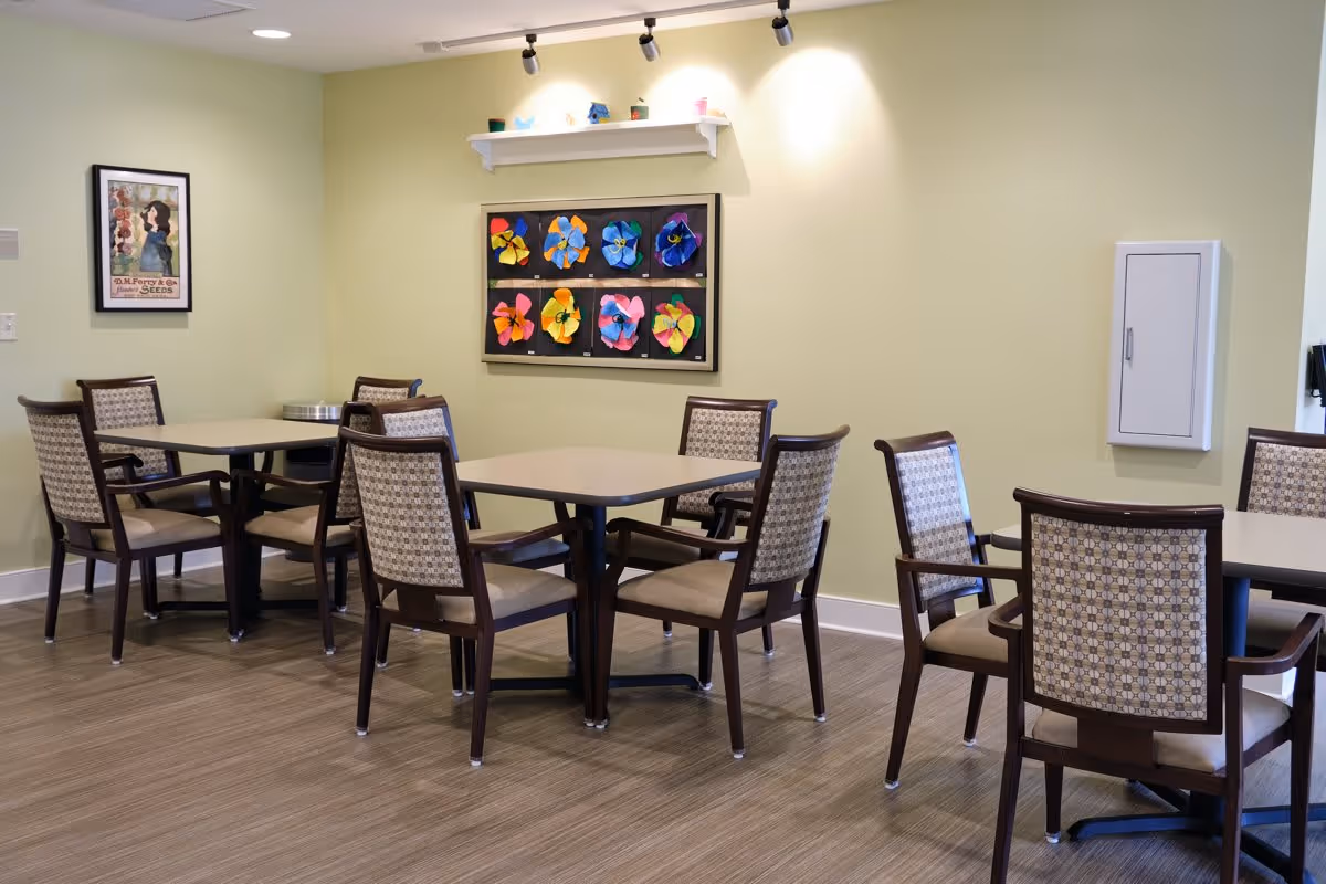 A dining area with several square tables and cushioned chairs arranged around them. The walls are painted light green and decorated with colorful flower artwork and a framed vintage-style poster. The floor has a wood-like finish, and there is a white wall-mounted cabinet on the right side.
