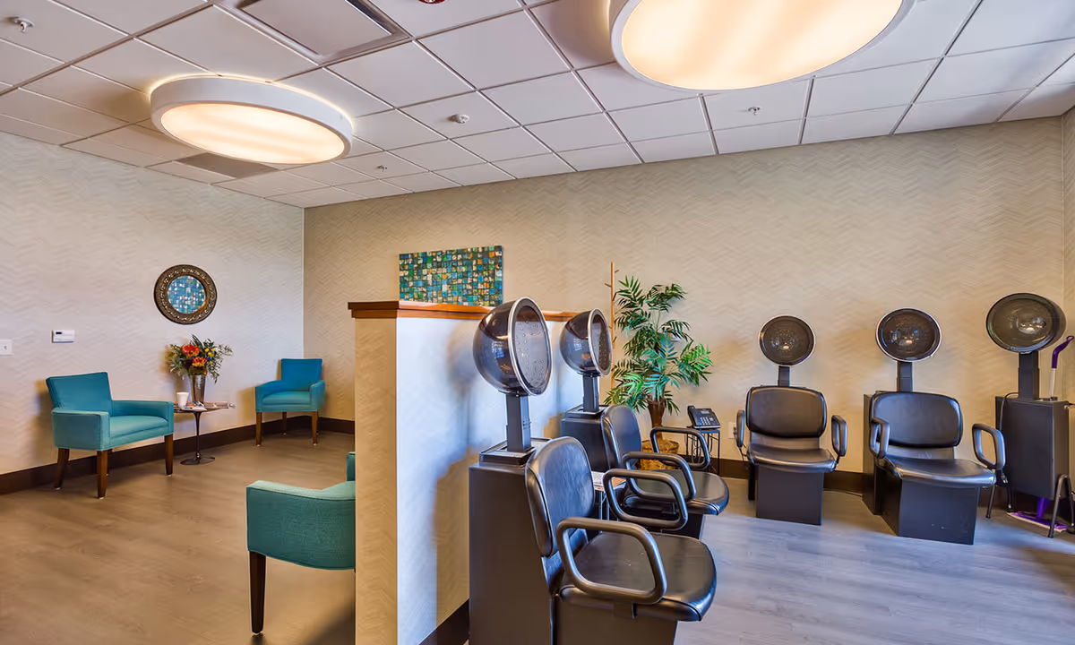 Interior view of a salon area in a senior living facility with multiple black salon chairs and hair dryers lined up against a wall. There are teal armchairs and a small table with flowers in the background. The room has a light-colored patterned wall and large round ceiling lights.