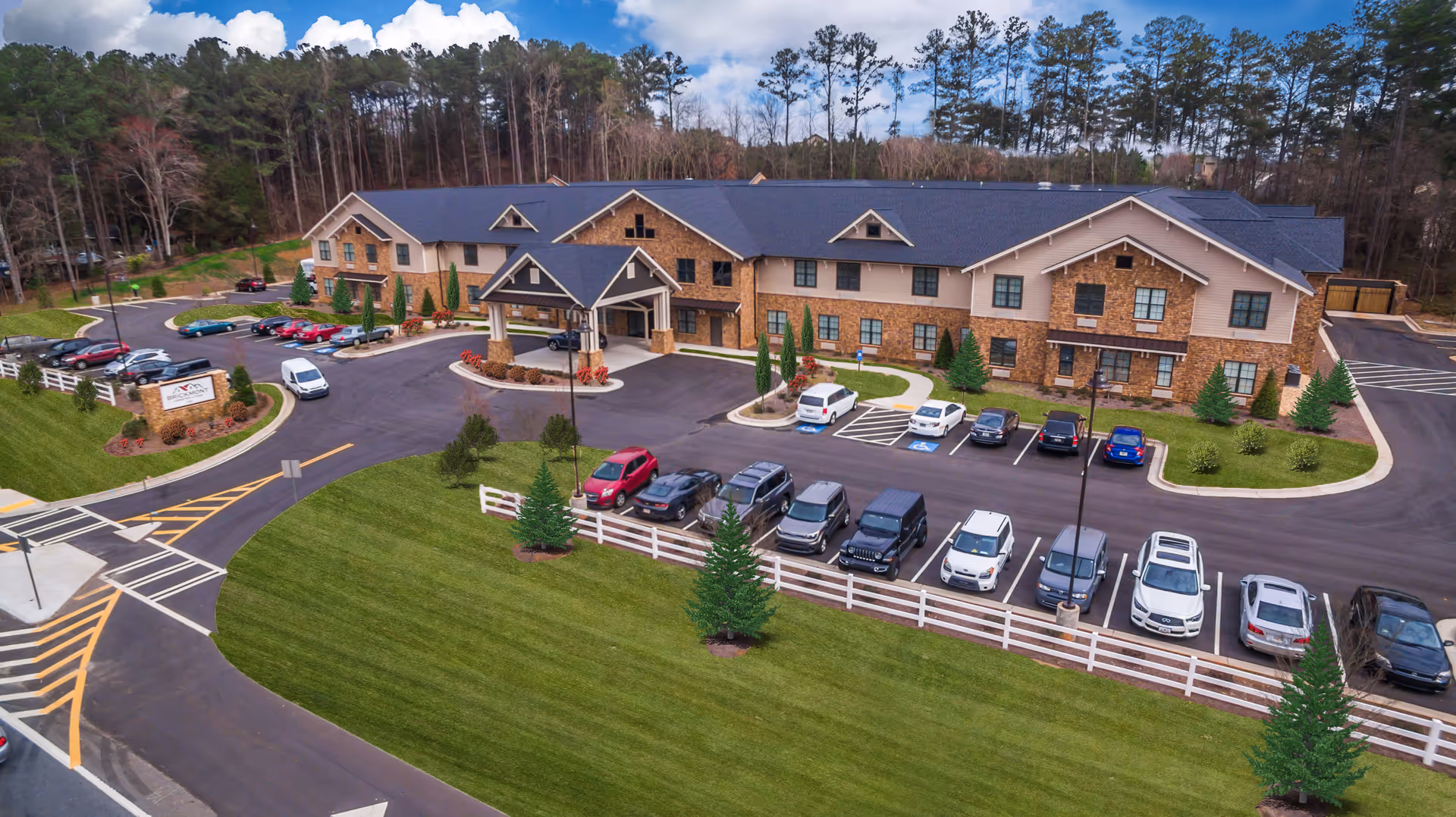 Aerial view of the Brickmont at Woodstock senior living building front with entrance canopy, parking lot, landscaped lawn, and surrounding trees.