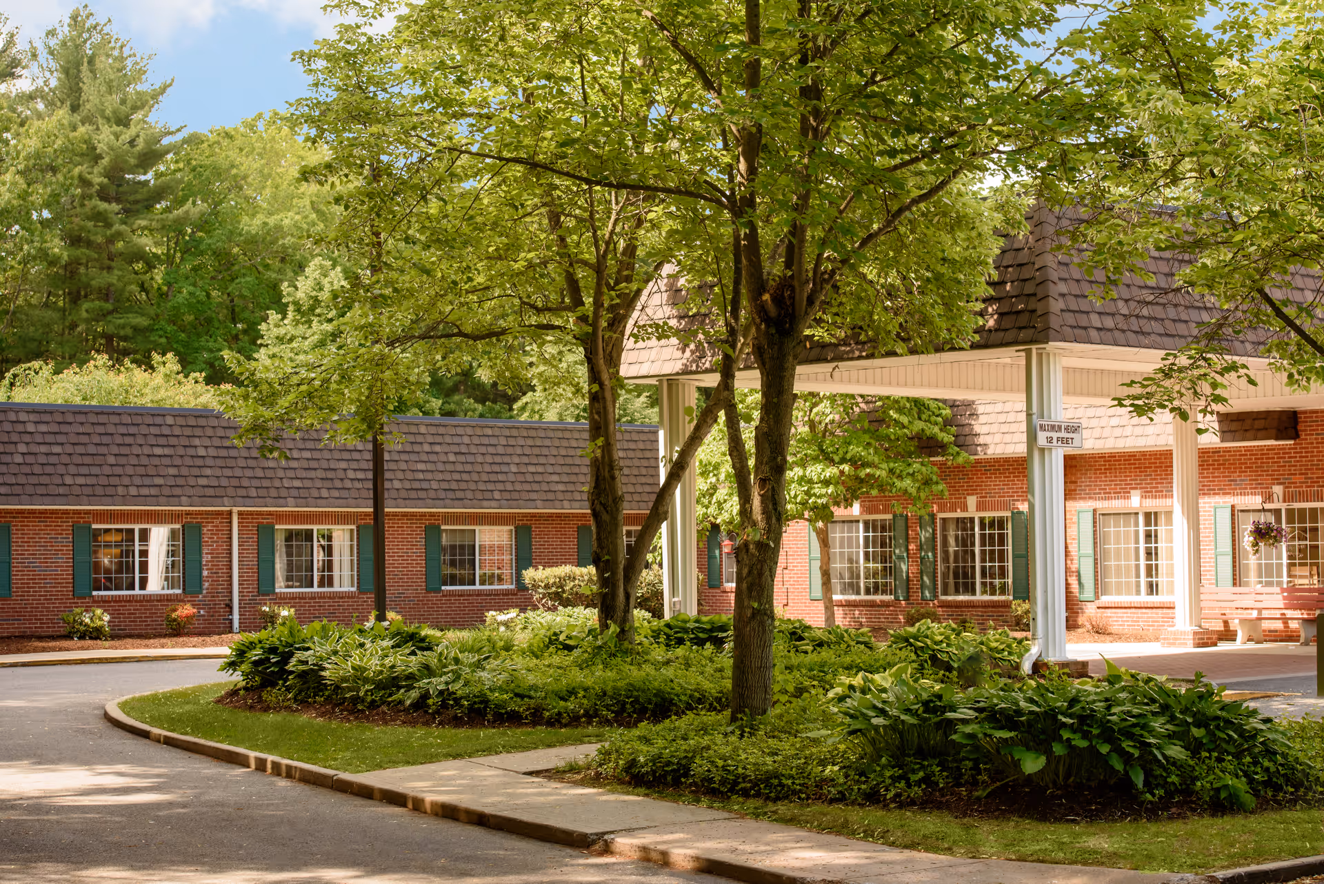 Front exterior of a brick senior living facility with a covered entrance, trees, and landscaped driveway.