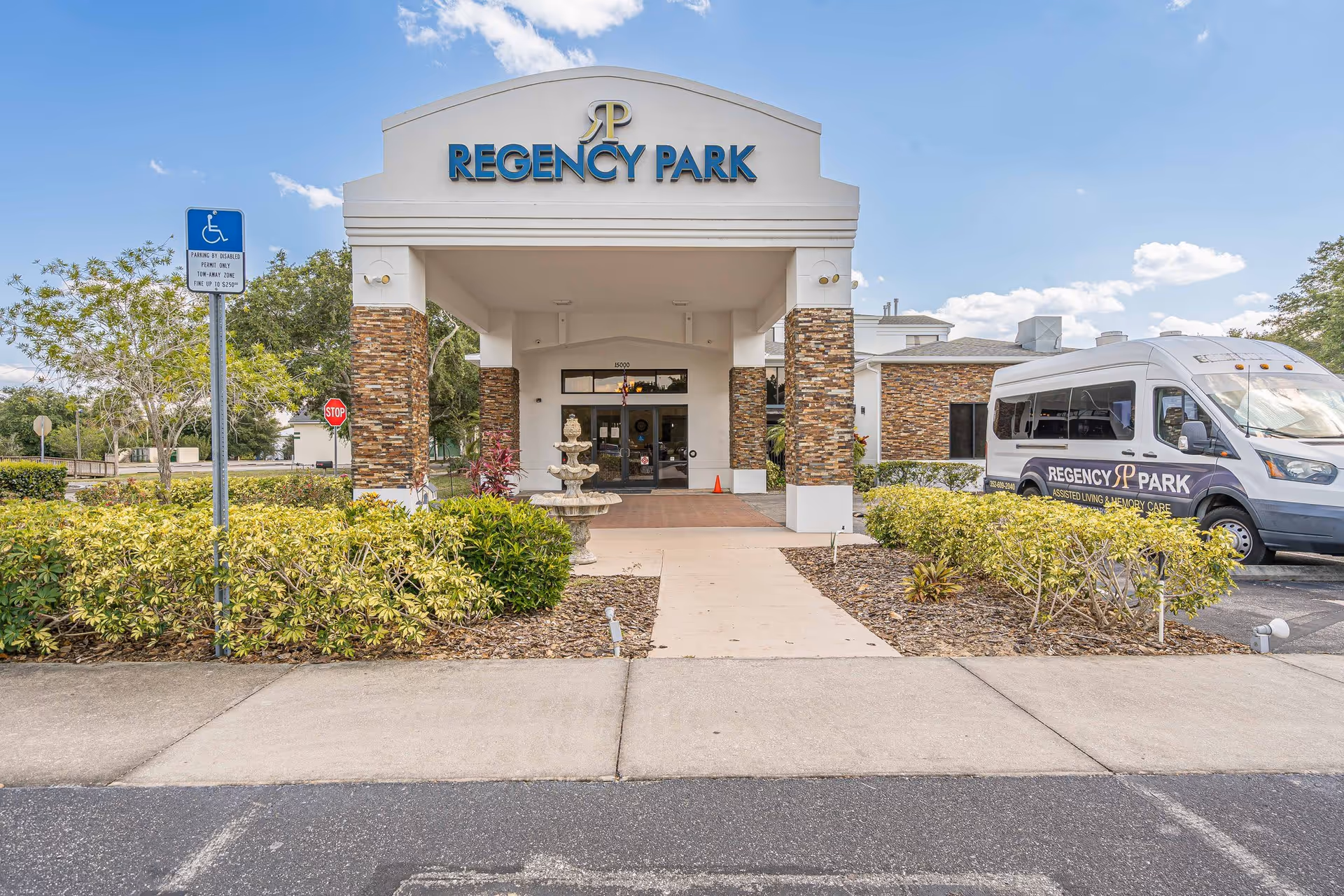 Front entrance of Regency Park Assisted Living & Memory Care facility with a covered walkway supported by stone pillars. There is a fountain in front of the entrance, surrounded by landscaped bushes. A Regency Park shuttle van is parked to the right. The sky is partly cloudy.