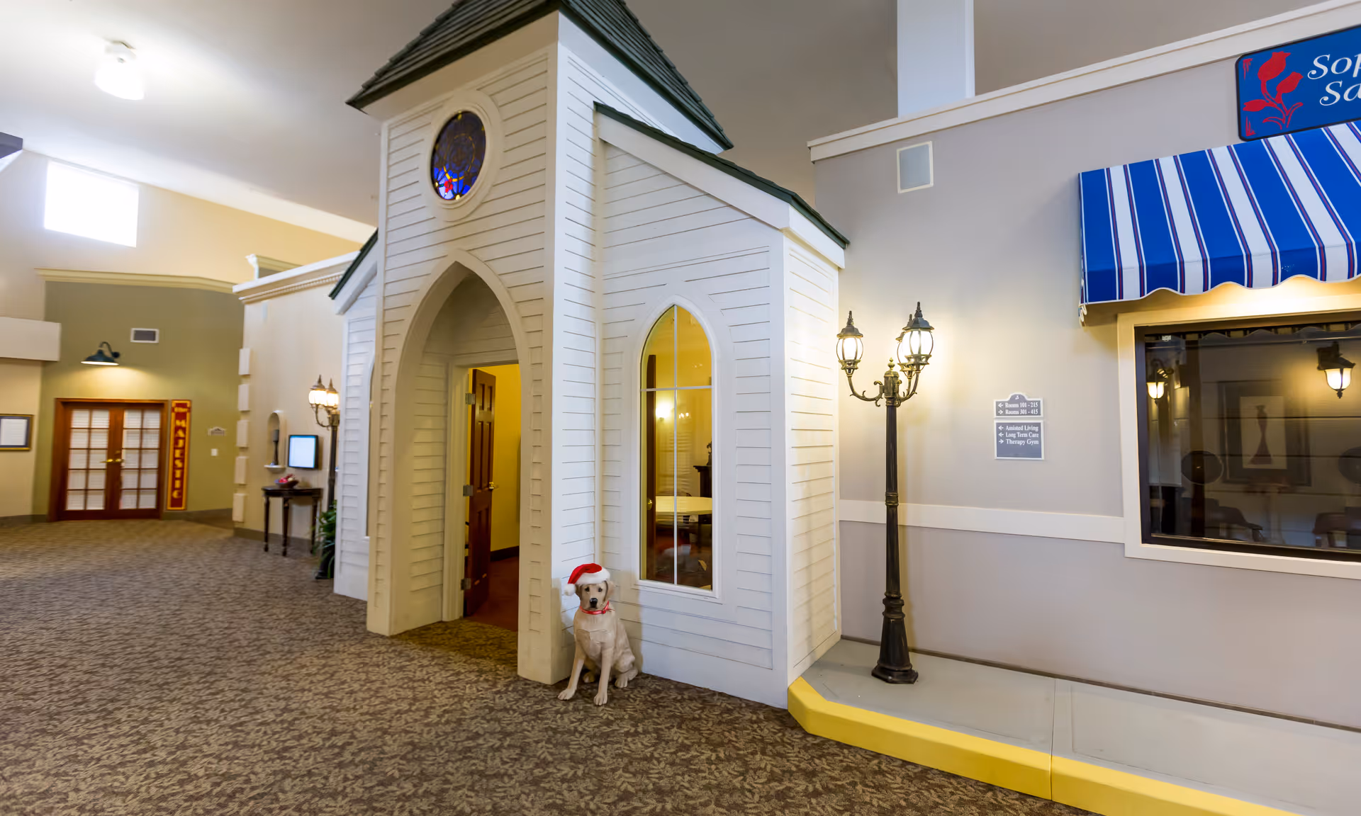 Interior of a senior living facility featuring a small white chapel-like structure with a stained glass window and an open door. A dog wearing a red Santa hat and collar sits at the entrance. To the right, there is a vintage-style street lamp and a blue and white striped awning above a window. The carpeted floor and walls are visible, along with directional signs for rooms and amenities.