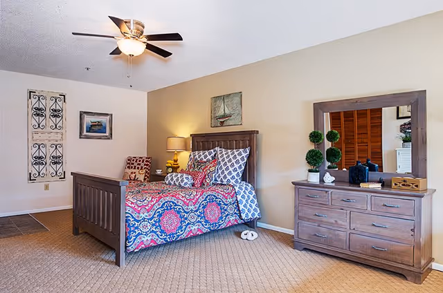 A cozy bedroom with a wooden bed frame and colorful patterned bedding. There is a nightstand with a lamp and a chair with a patterned cushion next to the bed. On the right side, there is a wooden dresser with a large mirror above it, decorated with small potted plants and other decorative items. The walls are light-colored, and the room has a ceiling fan with lights.