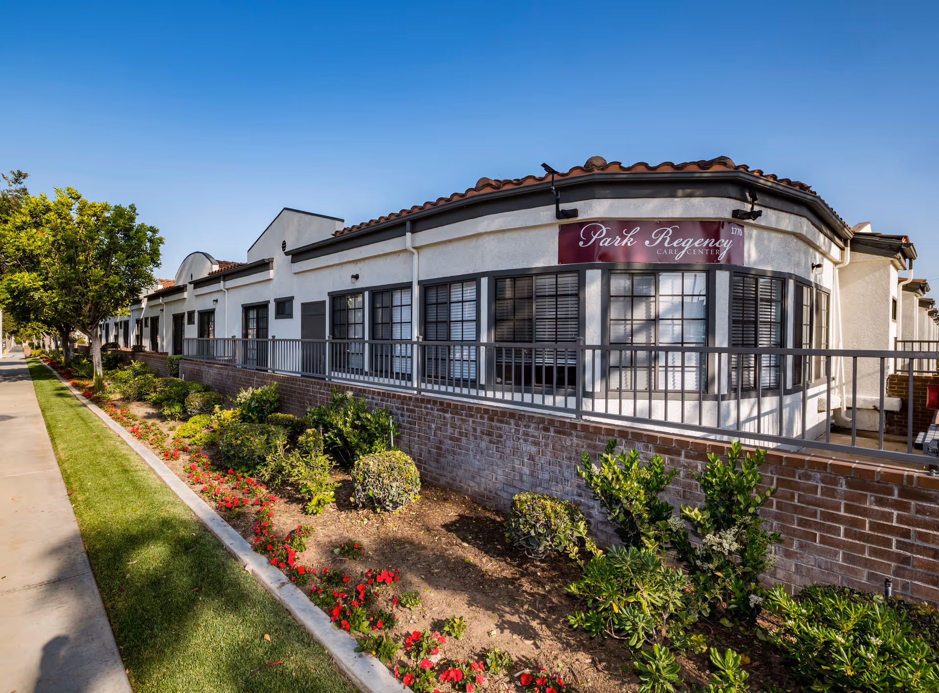 Exterior view of Park Regency Care Center building with white walls, large windows, and a red tiled roof. There is a garden with green shrubs and red flowers along the sidewalk in front of the building under a clear blue sky.