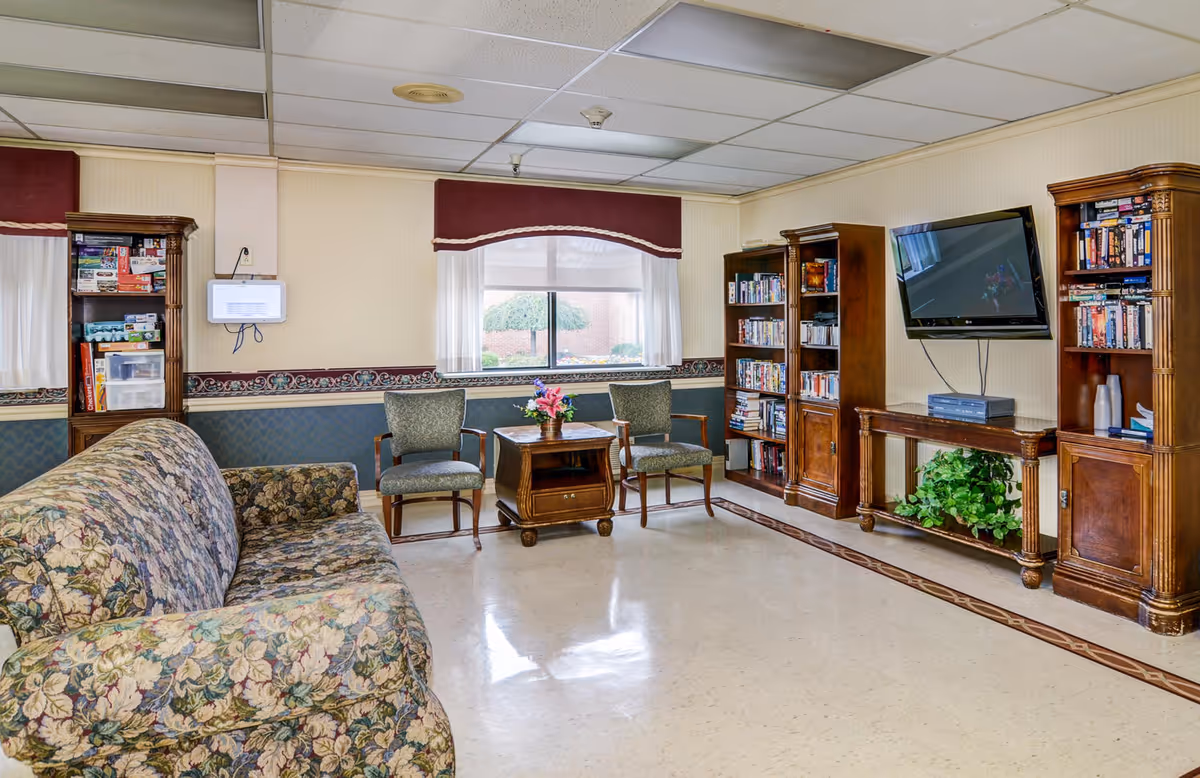 A cozy sitting area in a senior living facility featuring a floral couch, chairs, a coffee table, and shelves with books and movies.